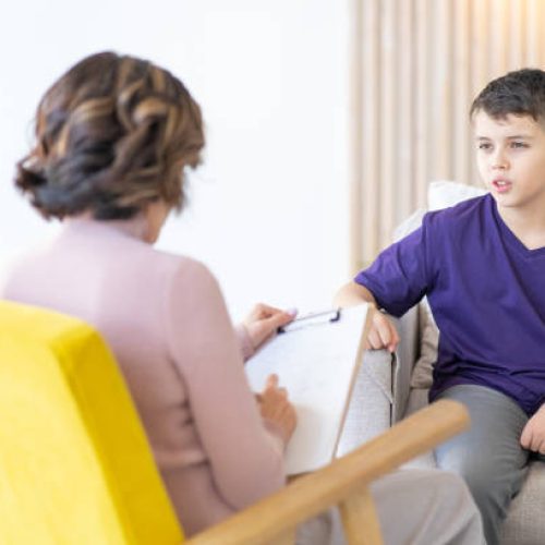 Teenage boy in purple T-shirt shares his experiences with psychotherapist. Woman psychotherapist in yellow chair in foreground blurred.